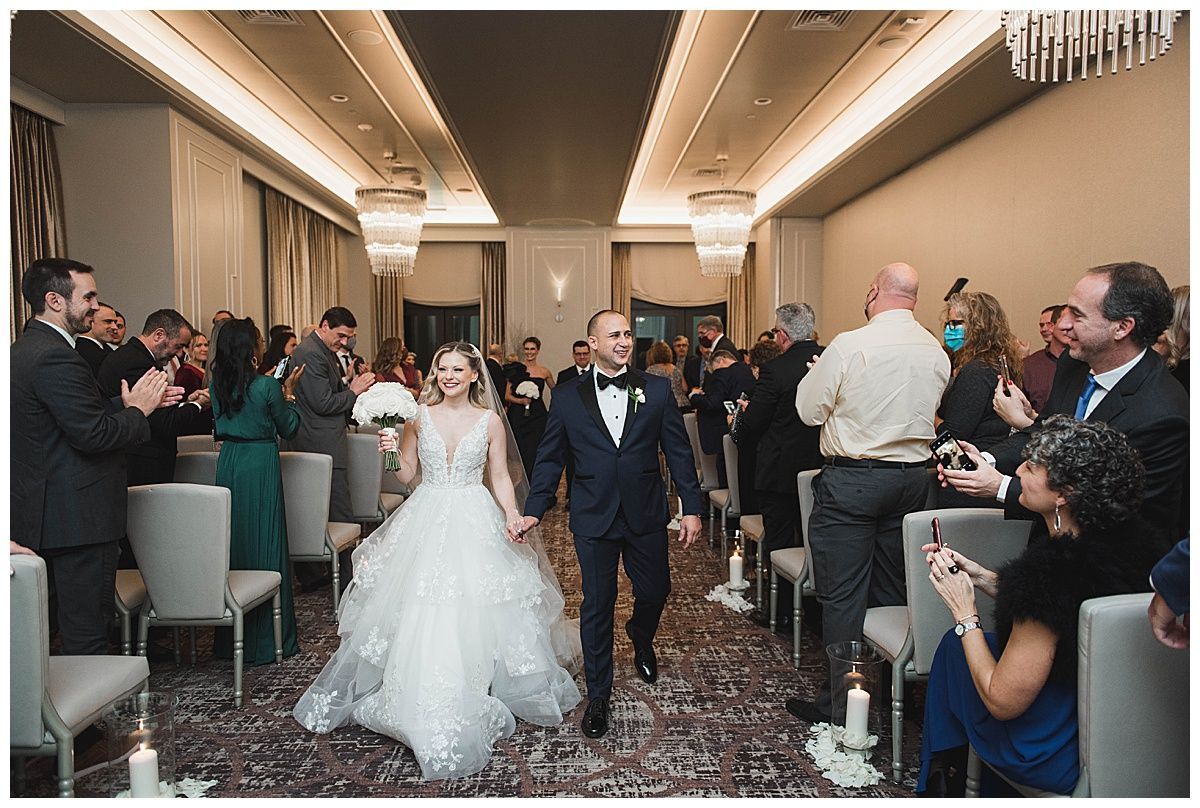 Newlyweds exiting wedding ceremony, holding hands, cheered on by guests seated in a decorated venue.