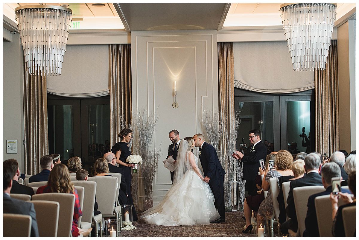 Wedding ceremony: Bride and groom kissing at altar, guests seated, chandeliers overhead.