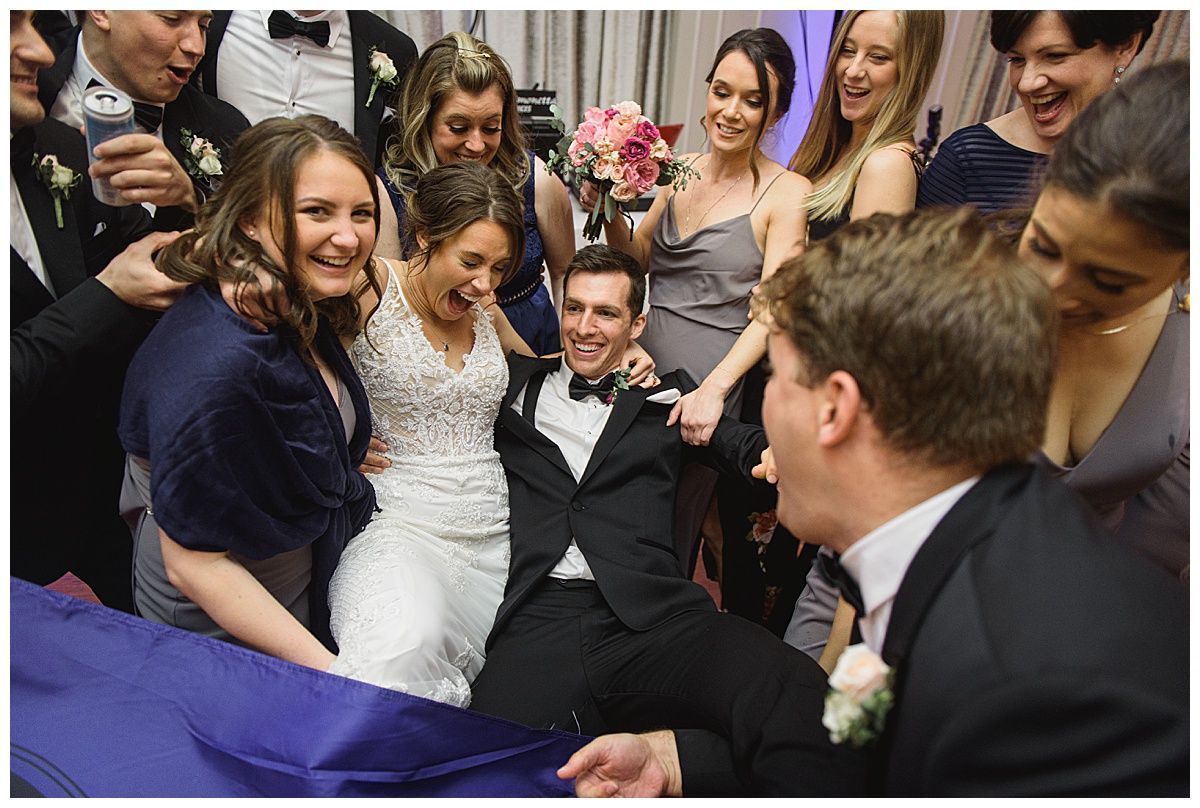 Wedding guests surround a laughing bride and groom, pulling them on a blue sheet.
