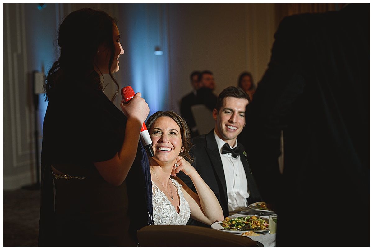 Woman giving a speech with a microphone; bride and groom seated, smiling at her, at a wedding reception.