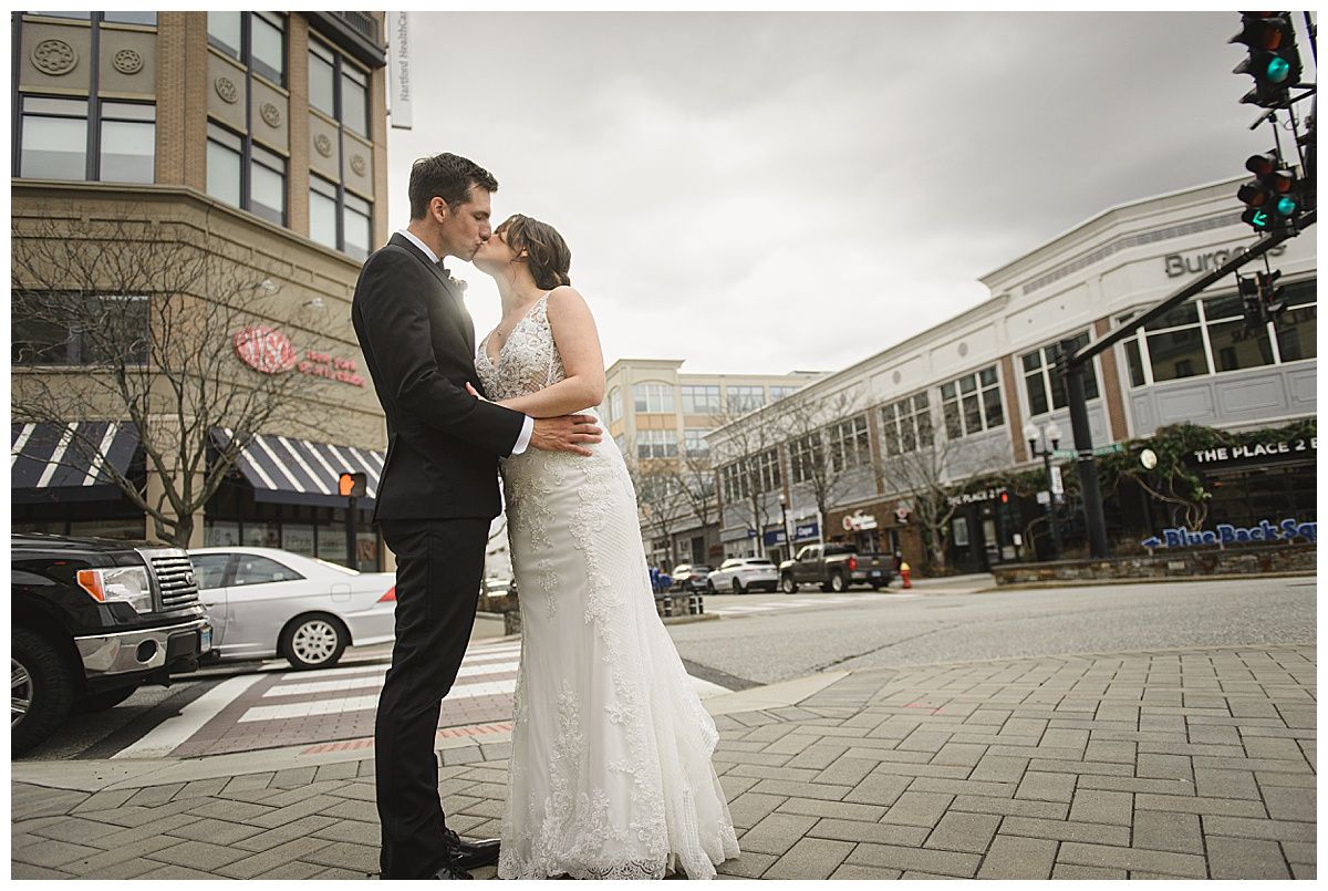 Bride and groom kissing in a city street. Bride in white lace dress, groom in black suit. Buildings in background.