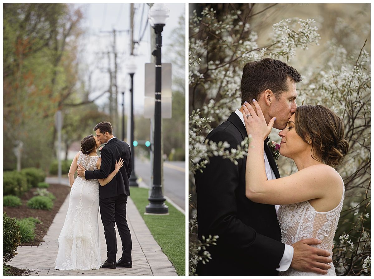 Wedding couple embraces; groom kisses bride's forehead. They're on a sidewalk and in front of flowering bushes.