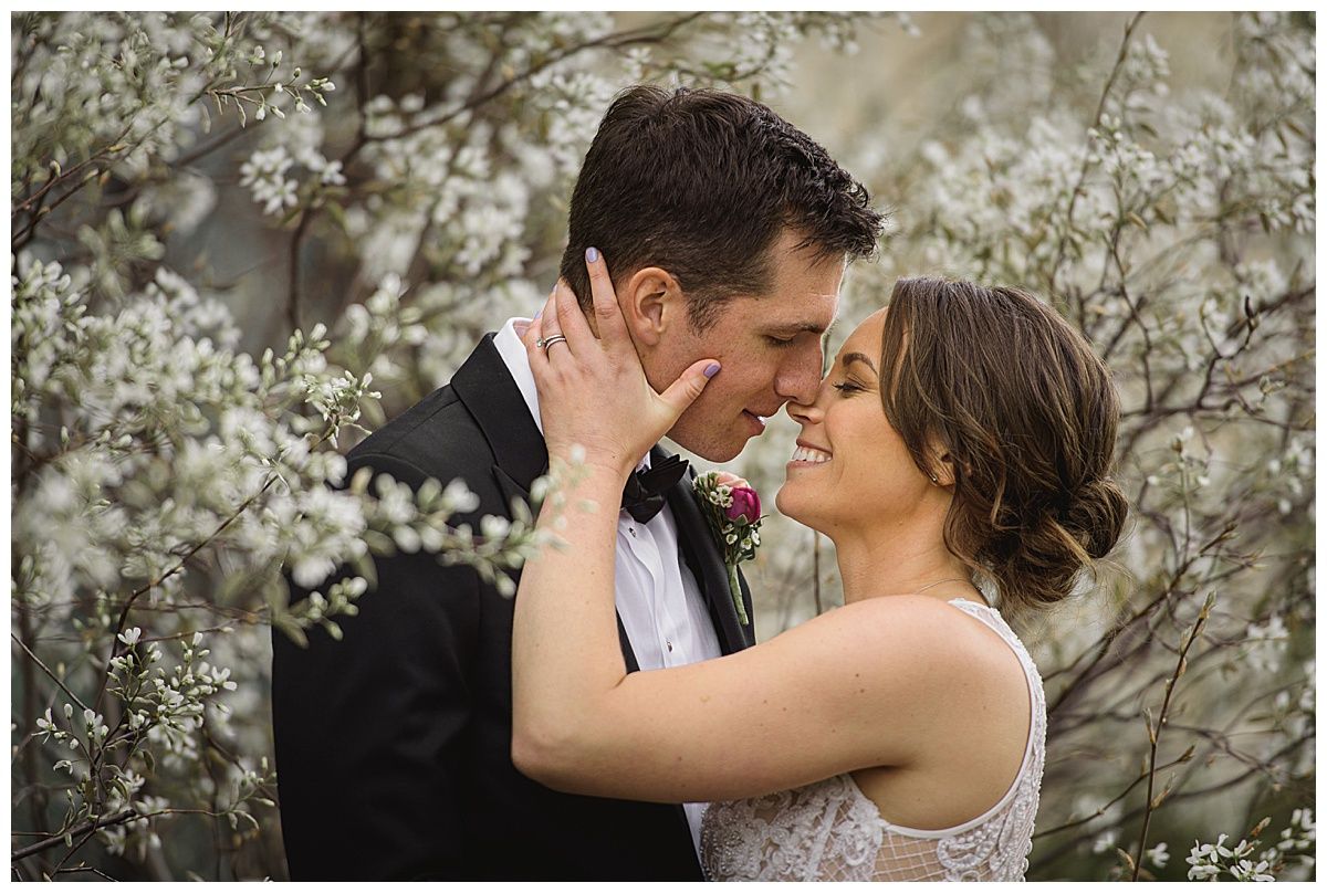 Couple embraces, nearly kissing, surrounded by white flowers. The man wears a tuxedo. The woman wears a white dress.