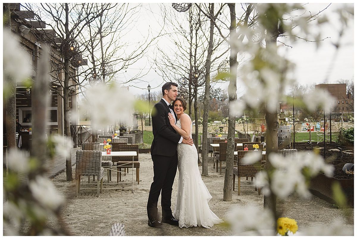Wedding couple embraces outdoors, framed by white flowers; dining tables visible.