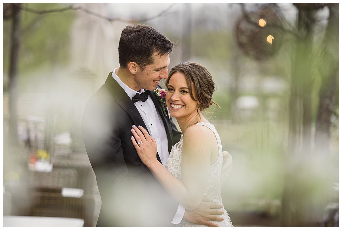 Bride and groom embrace, smiling. The groom wears a black tuxedo. The bride wears a white wedding dress. Outdoor setting.