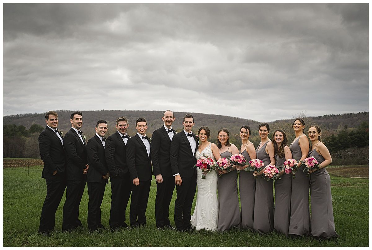 Wedding party posing in a field. Bridesmaids in gray dresses hold pink bouquets, groomsmen in black tuxedos. Overcast sky.