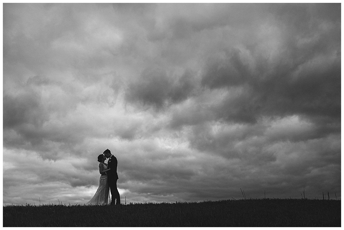 Couple kissing under a dramatic, cloudy sky. Silhouetted on a grassy horizon.