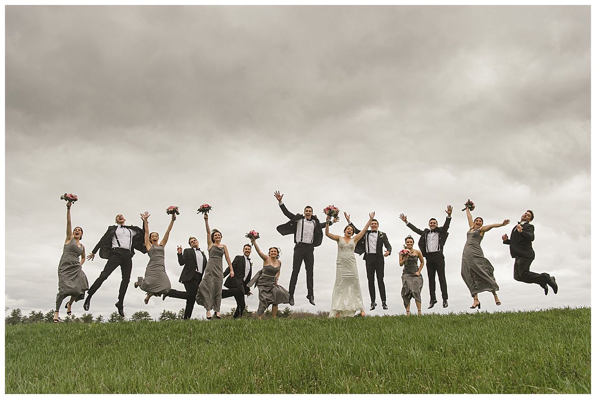 Wedding party jumping on a grassy hill under a cloudy sky; dresses are gray, suits are black.