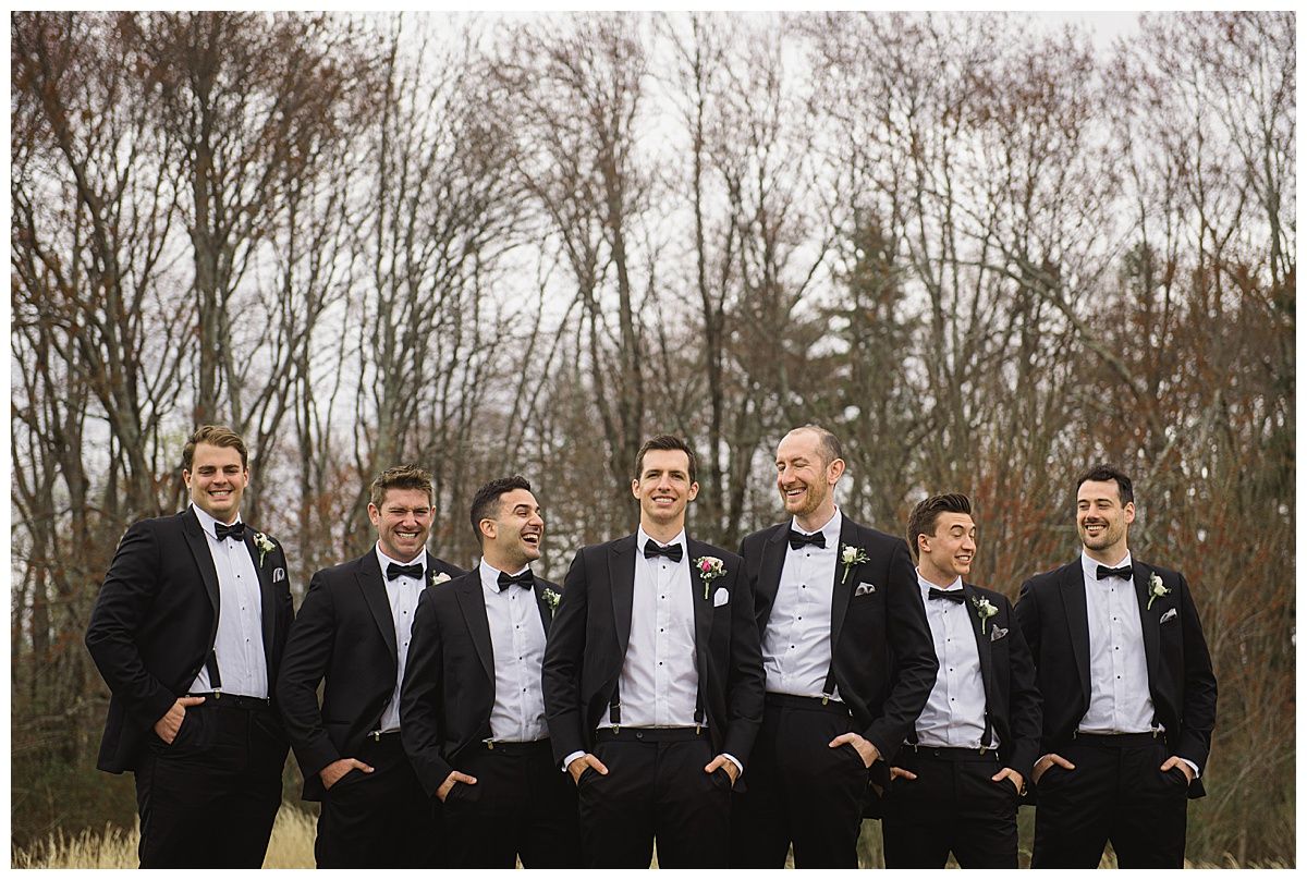 Seven men in tuxedos, smiling, posing together outdoors with bare trees in the background.