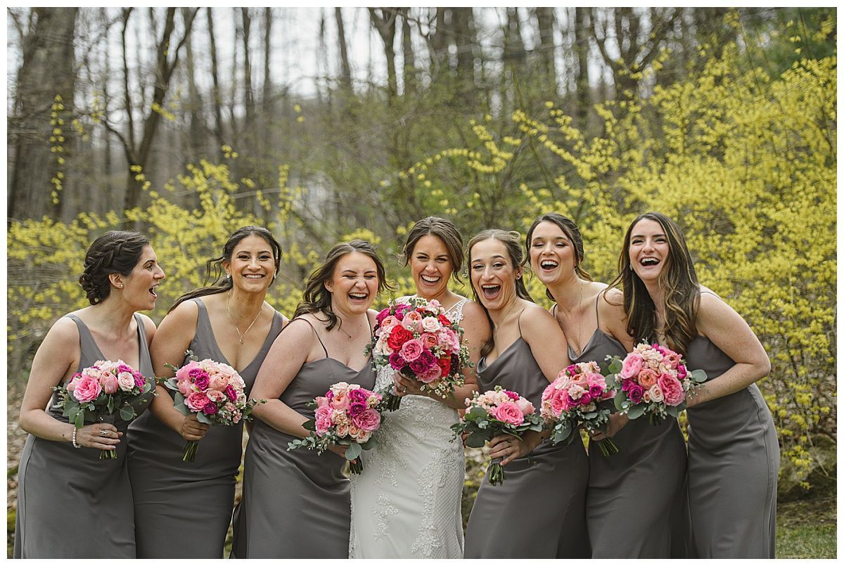 Bride with bridesmaids laughing, holding bouquets, wearing gray dresses outdoors near yellow flowering bushes.
