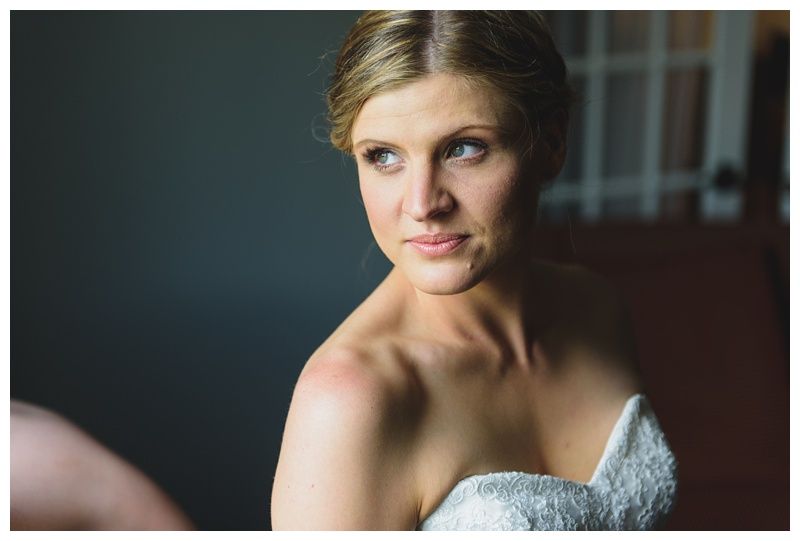 Bride in white gown holding bouquet, posed outdoors. Sunlight streams through trees.