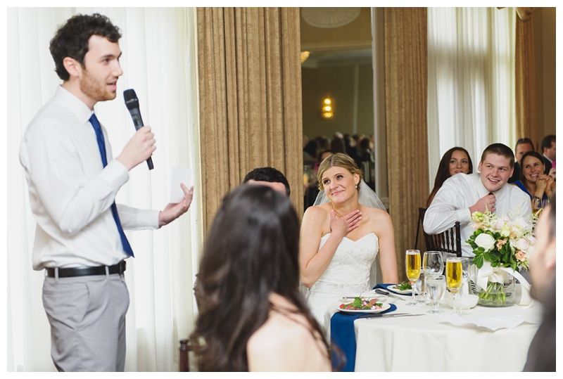 Bride with arms up, making a funny face, groom looking at his hand, cake cutting in background.