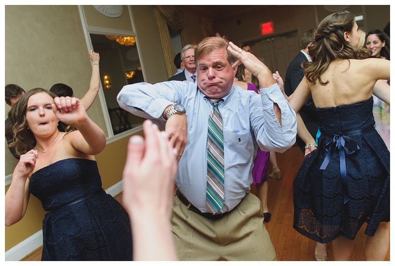 Bride with arms up, making a funny face, groom looking at his hand, cake cutting in background.