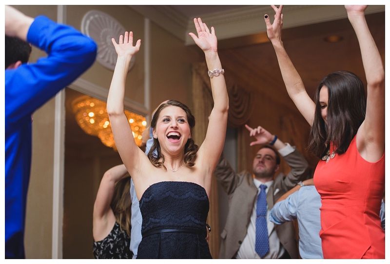 Bride with arms up, making a funny face, groom looking at his hand, cake cutting in background.