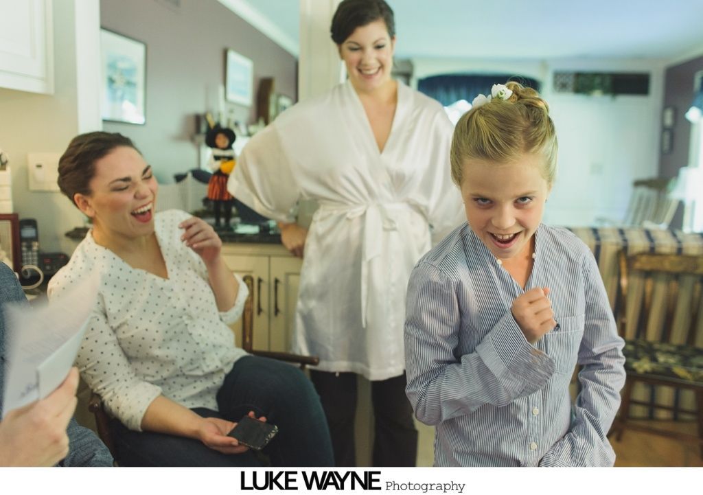 A young person excitedly grins, arm raised, with two women laughing in a room with furniture.