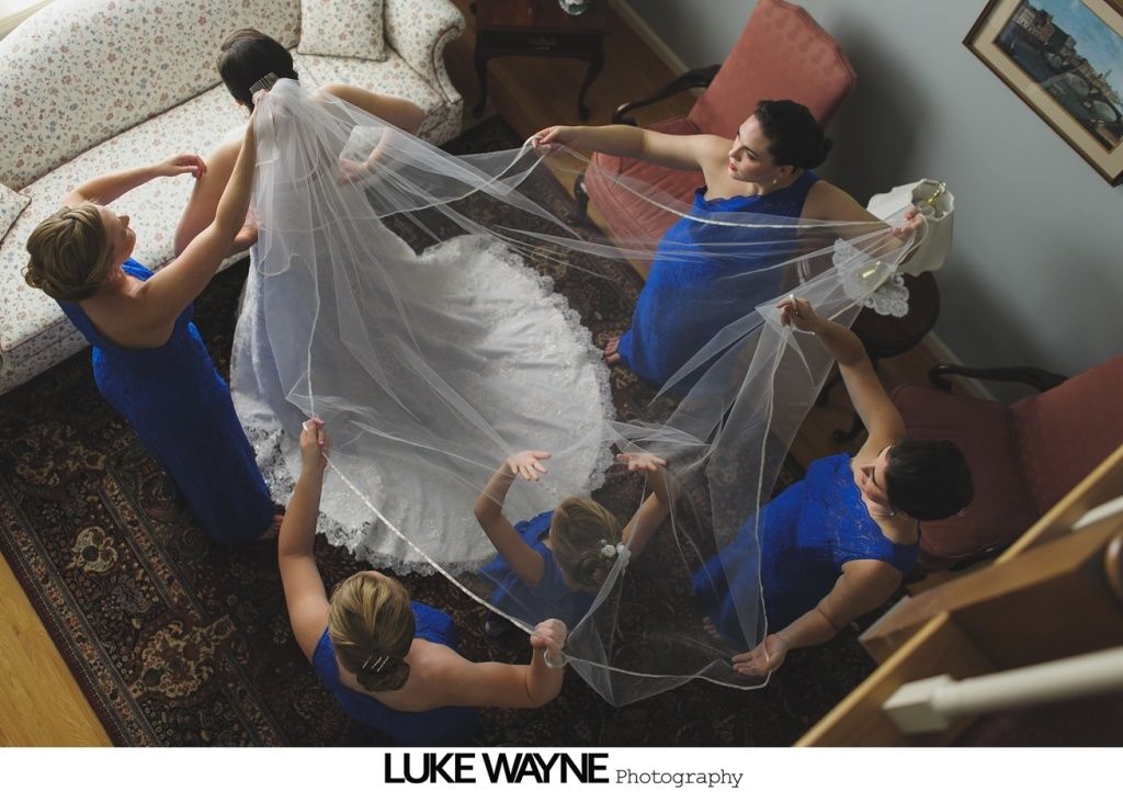 Bride surrounded by bridesmaids holding veil, wearing blue dresses, inside a house.