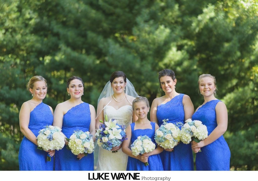 Bride and bridesmaids in blue dresses with bouquets, posed in front of a green tree backdrop.