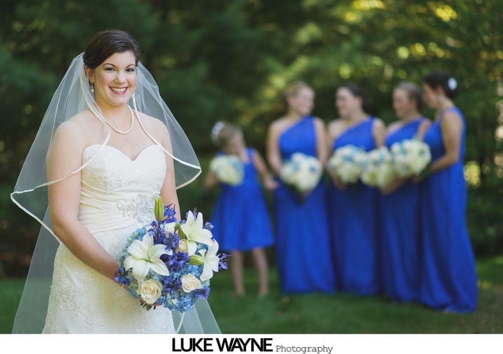 Bride in white dress holding flowers, smiles, with bridesmaids in blue dresses in a garden setting.