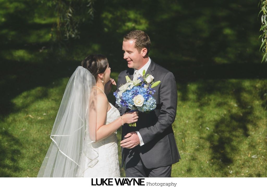 Bride and groom smiling at each other, holding bouquet, in a sunlit garden.