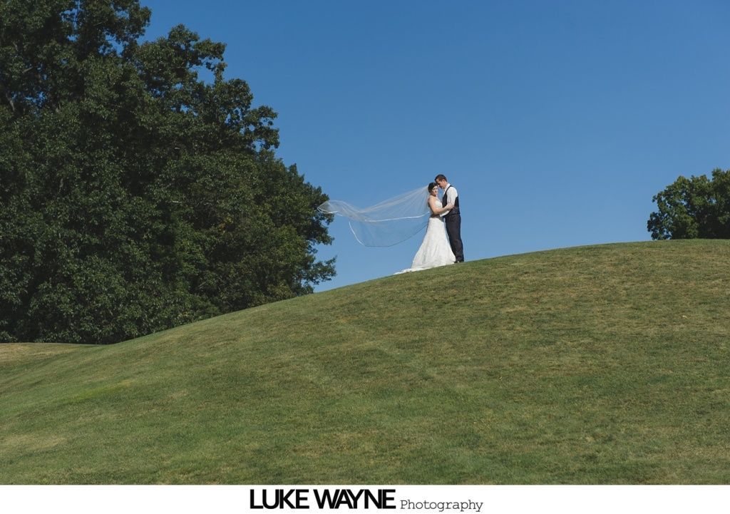 Bride and groom kissing on a grassy hill; veil blowing in the wind, blue sky.