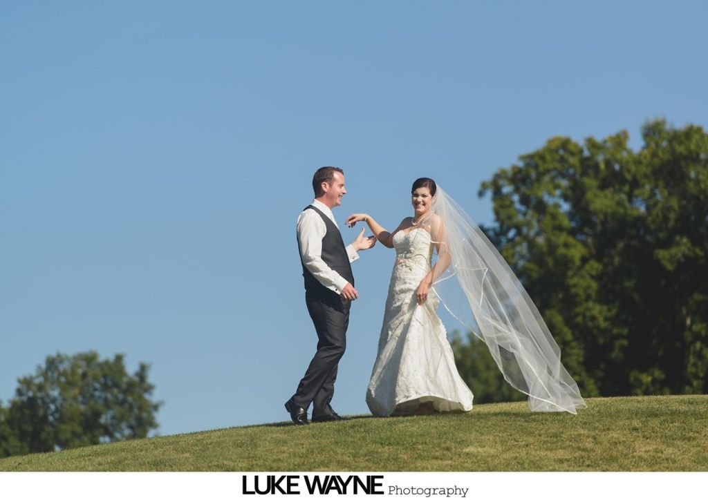Bride and groom on a grassy hill, dancing, blue sky, trees in background.