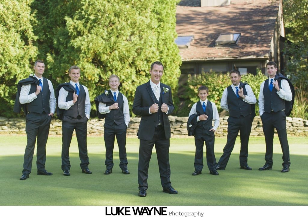 Groom and groomsmen in black suits, blue ties, on green grass. Building in background, all smiling.