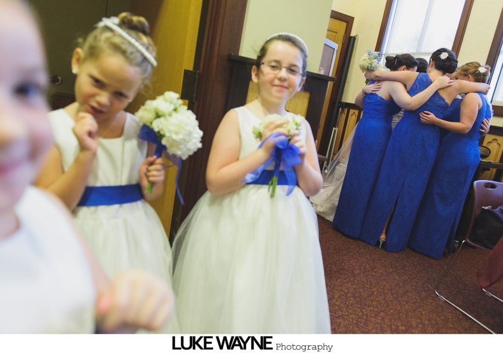 Girls in white dresses with blue sashes hold bouquets, looking at camera. Bridesmaids in blue dresses embrace.