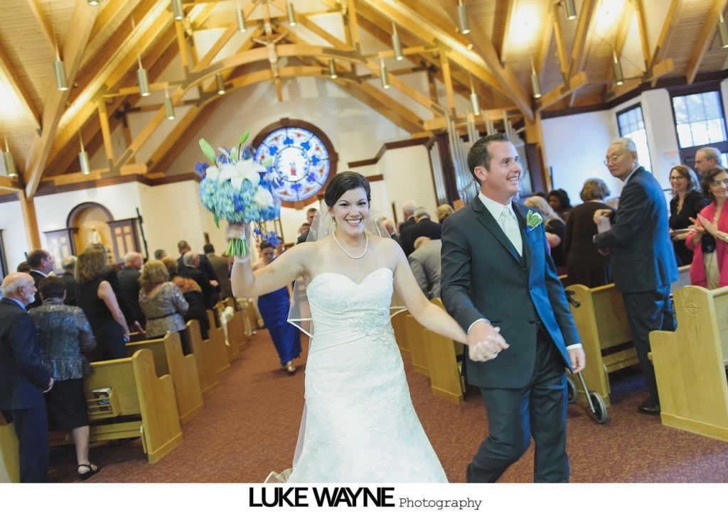 Newlyweds exit a church, smiling, holding hands. Bride in white gown, groom in suit. Guests clap.
