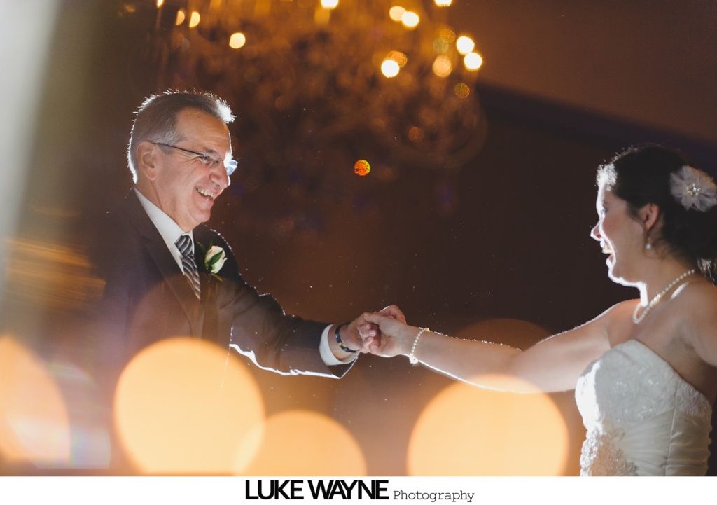 Bride and father dance, holding hands, smiling. Warm lighting in a room with a chandelier.