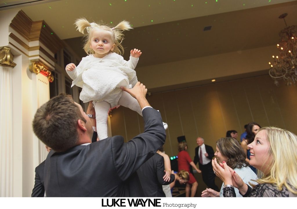 Man tossing a young child with blonde pigtails in the air at a party, other people clapping.