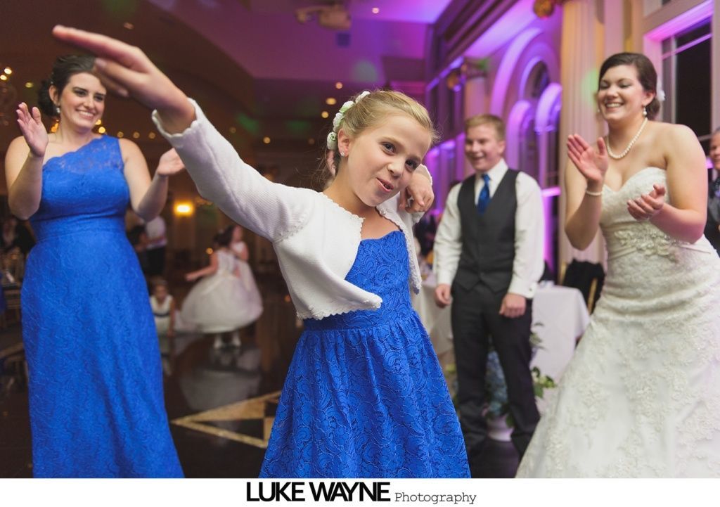 Girl dabs on a dance floor, flanked by a woman in a blue dress and a bride.