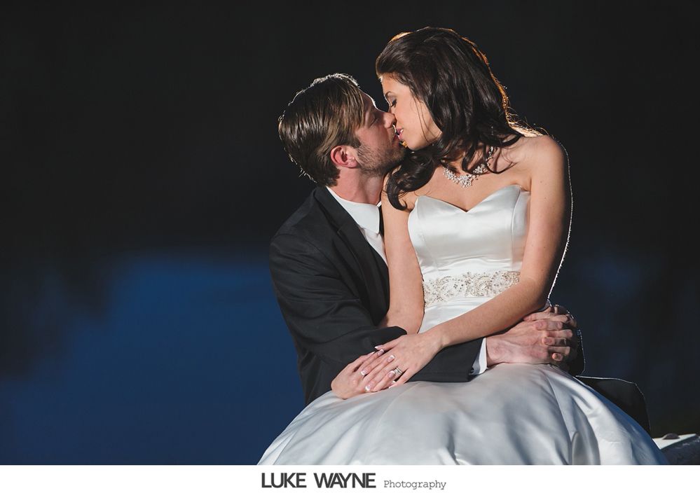 Bride and groom embrace in a kiss; she wears a white gown, he a black suit, with a dark background.