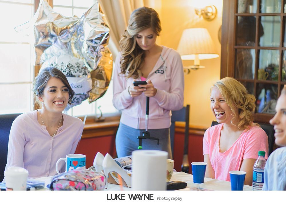 Women in pink shirts at a table, some laughing. One holds a phone on a stick. Balloons, shoes, and cups on the table.