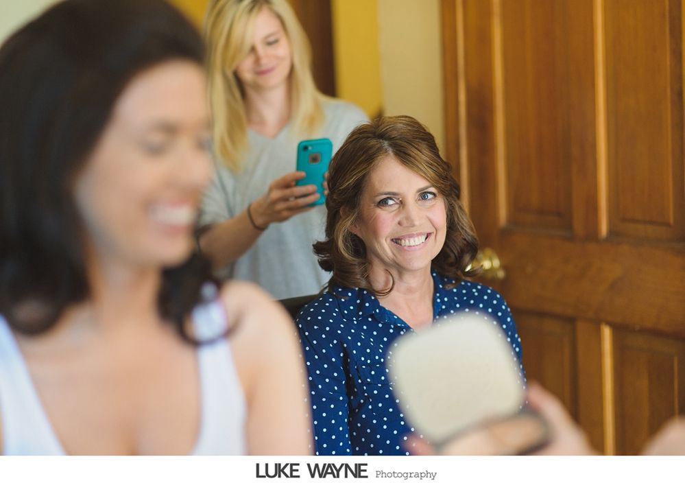 Woman in blue polka-dot shirt smiles, holding a mirror, as another takes a photo in front of a wood door.