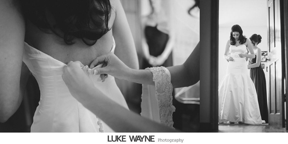 Bride getting dressed, back view, with assistance from another person. Mirror reflection shows the bride and bridesmaid.