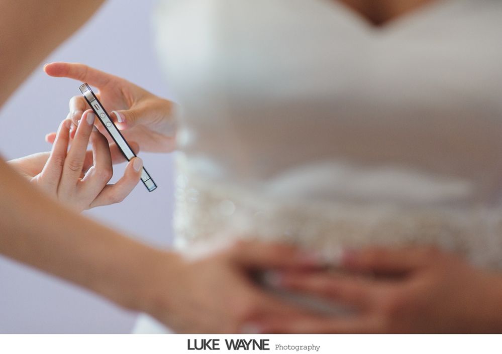 Bride holding a phone; another person assisting with the wedding dress.