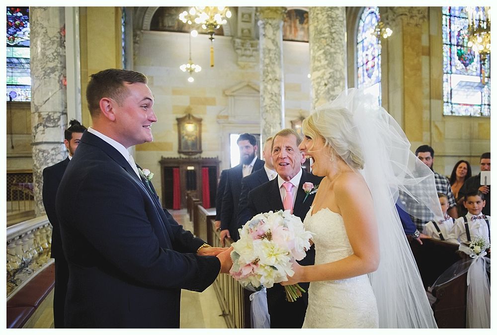 Bride with arms up, making a funny face, groom looking at his hand, cake cutting in background.