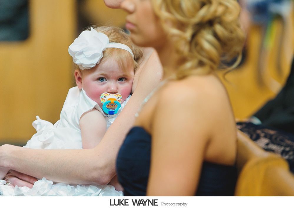 Baby in white dress with pacifier, flower headband, held by woman in a black strapless dress.