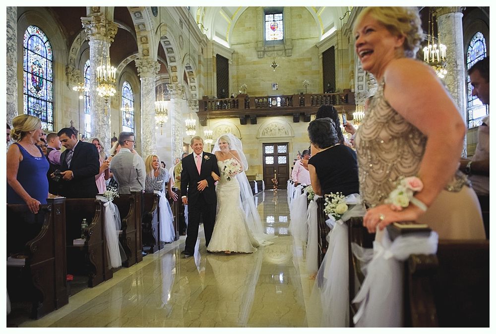 Bride with arms up, making a funny face, groom looking at his hand, cake cutting in background.