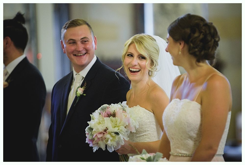 Bride with arms up, making a funny face, groom looking at his hand, cake cutting in background.