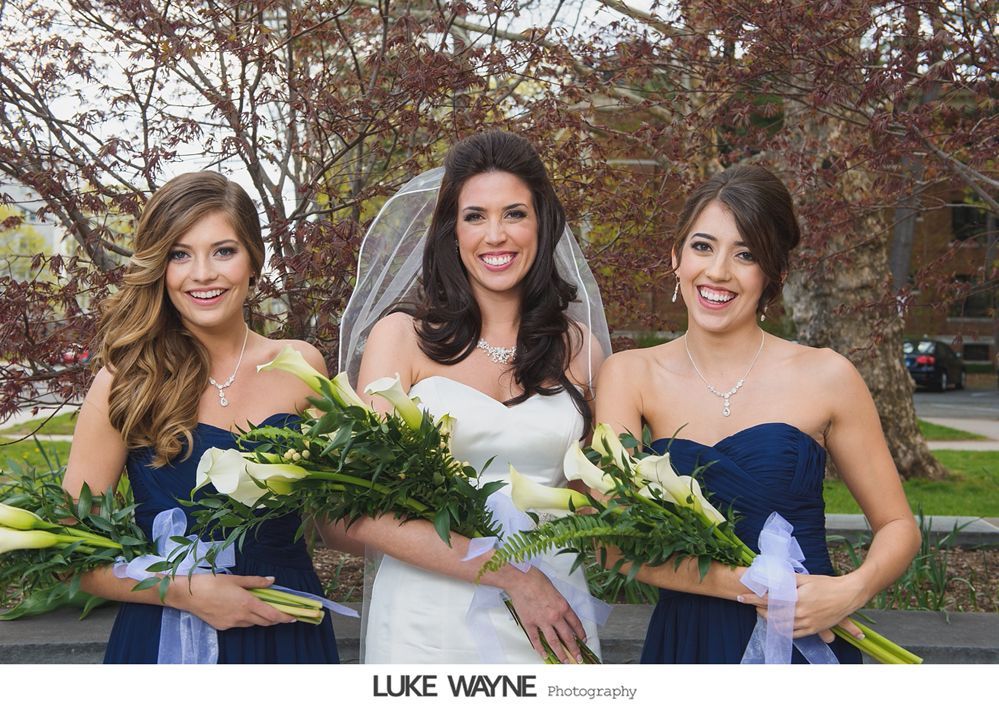 Bride and bridesmaids in navy blue dresses holding calla lily bouquets, smiling. Outdoor setting.