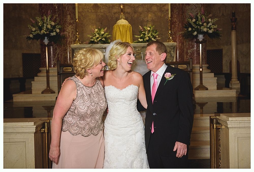 Bride with arms up, making a funny face, groom looking at his hand, cake cutting in background.