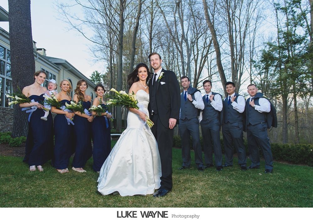 Wedding party: Bride and groom stand with bridesmaids in blue dresses and groomsmen in grey suits, outside a building.