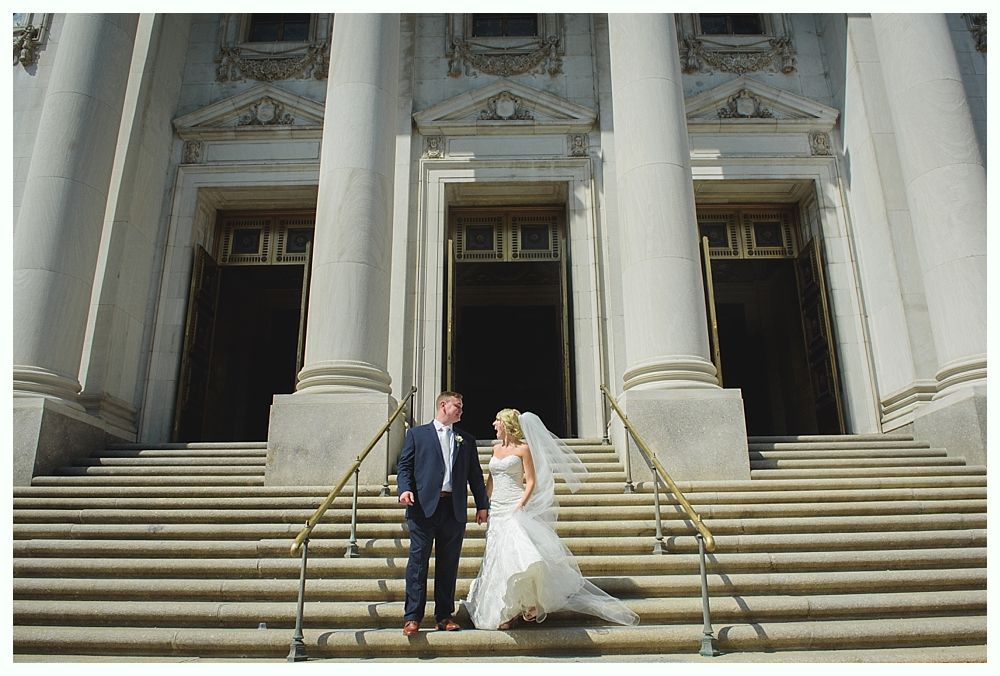 Bride with arms up, making a funny face, groom looking at his hand, cake cutting in background.