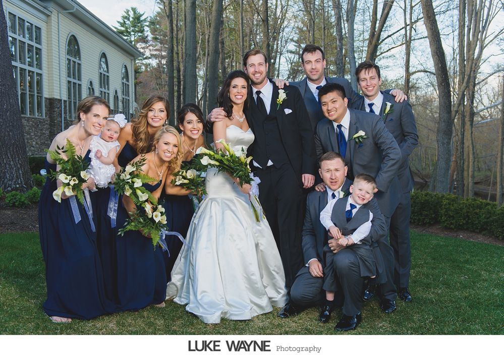 Wedding party poses outdoors; bride in white gown, groom in black suit, bridesmaids in navy, groomsmen in gray.