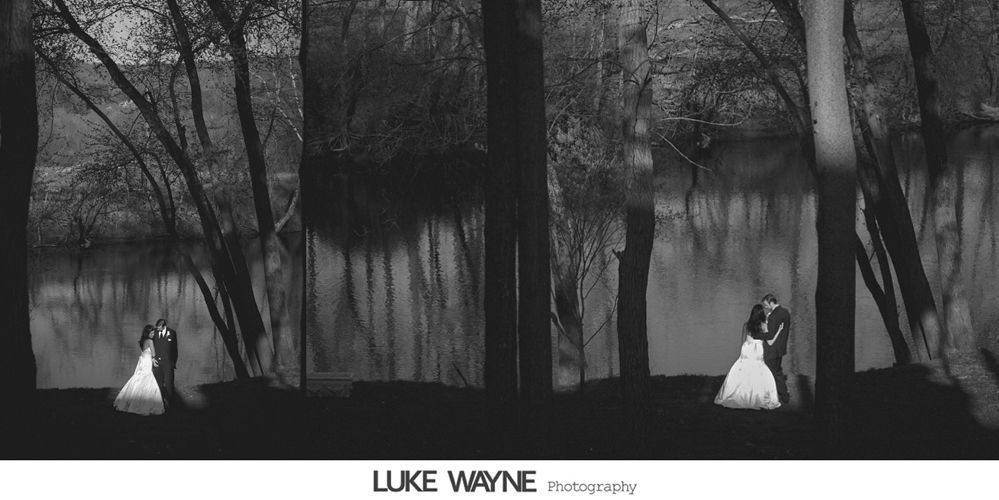 Black and white photo of a couple in wedding attire by a lake, standing between trees.