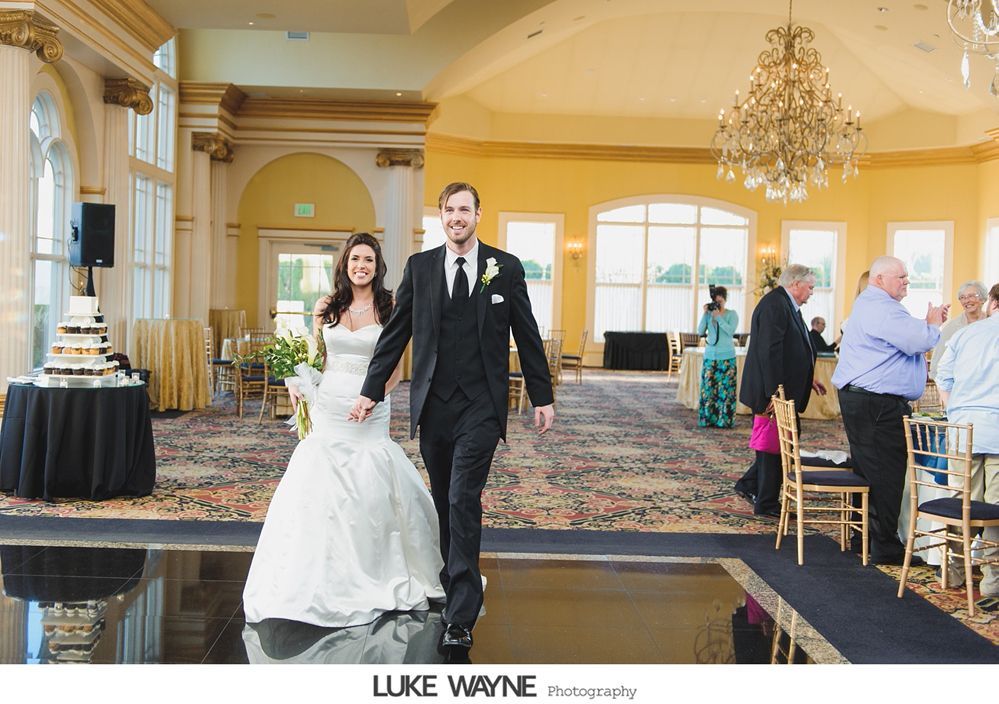 Newlyweds walk hand-in-hand across a dance floor in a ballroom, smiling.
