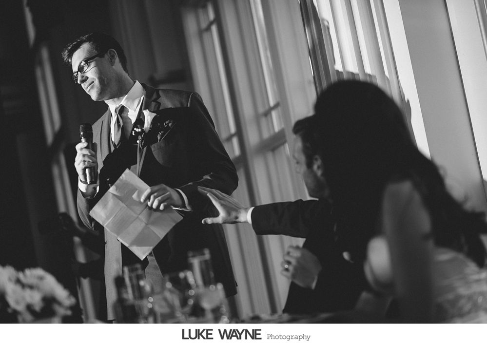 Man in suit gives a speech with a microphone, gesturing towards seated guests in a well-lit room.