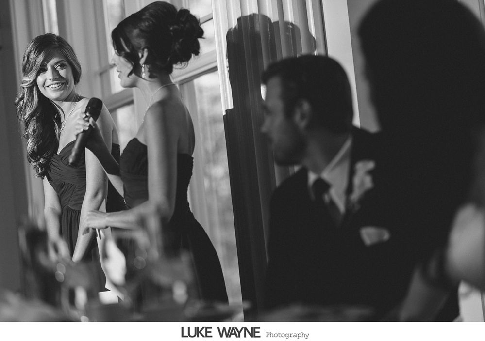Woman giving a speech at an event, smiling, holding a microphone. Other people seated at a table.