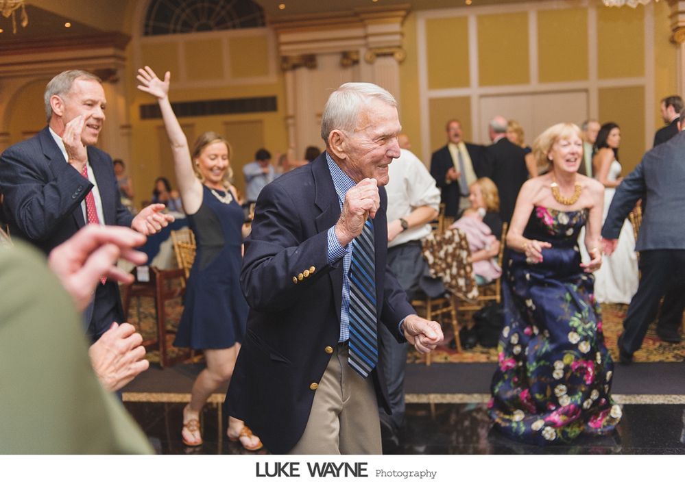 People dancing at a wedding reception. Elderly man in a blue blazer dances with a smile. Others also dance.